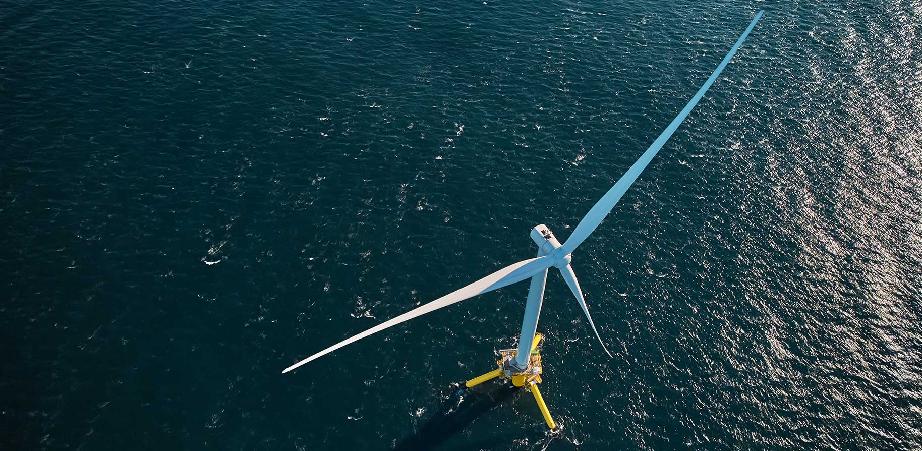 An aerial view of a large white wind turbine standing tall on a yellow platform in the deep blue sea.