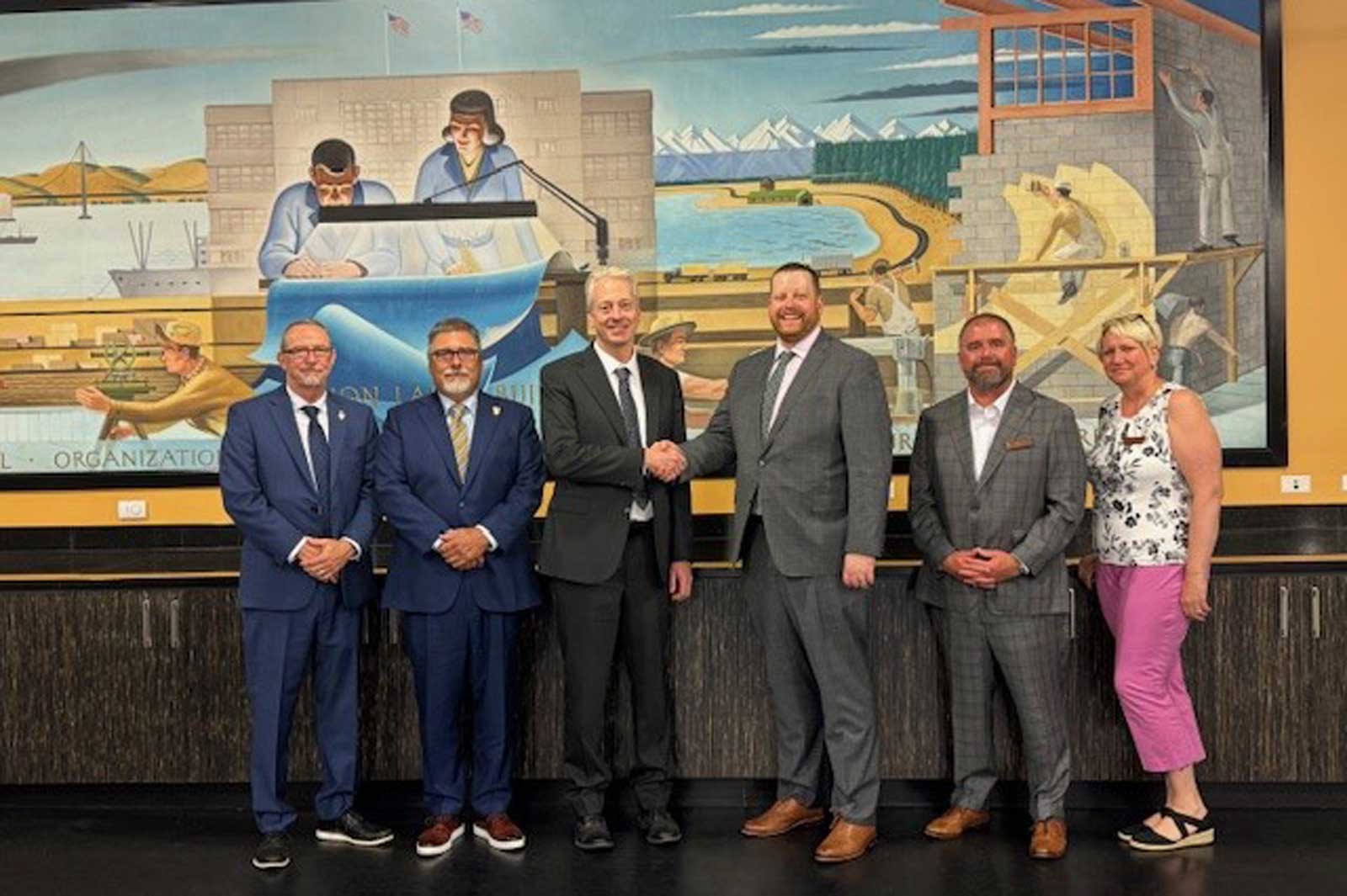 A group of five business people stands in front of a mural depicting maritime and construction scenes. They are all in formal attire.