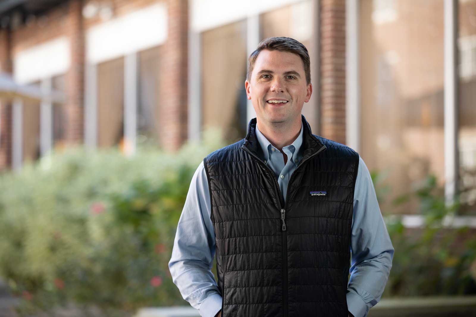 A person in a black vest over a light blue shirt stands outdoors near plants and brick buildings.
